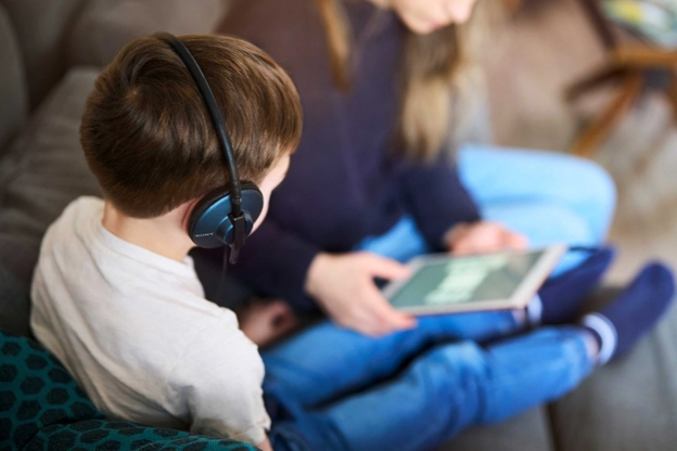 Kind sitzt mit Kopfhörern und Teddybär mit seiner Mutter am Tablet